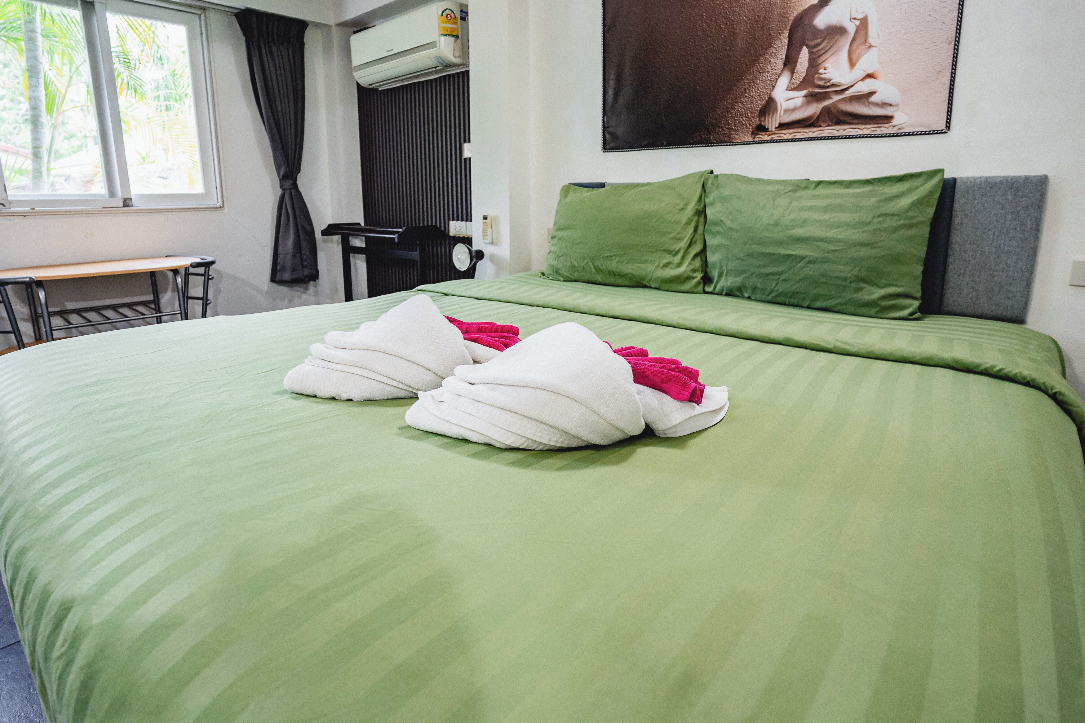Hotelkamer met een groen dekbedovertrek, twee witte handdoeken, een groen kussen, een grijs kussen, een zwarte stoel, een bureau en een raam met gordijnen.