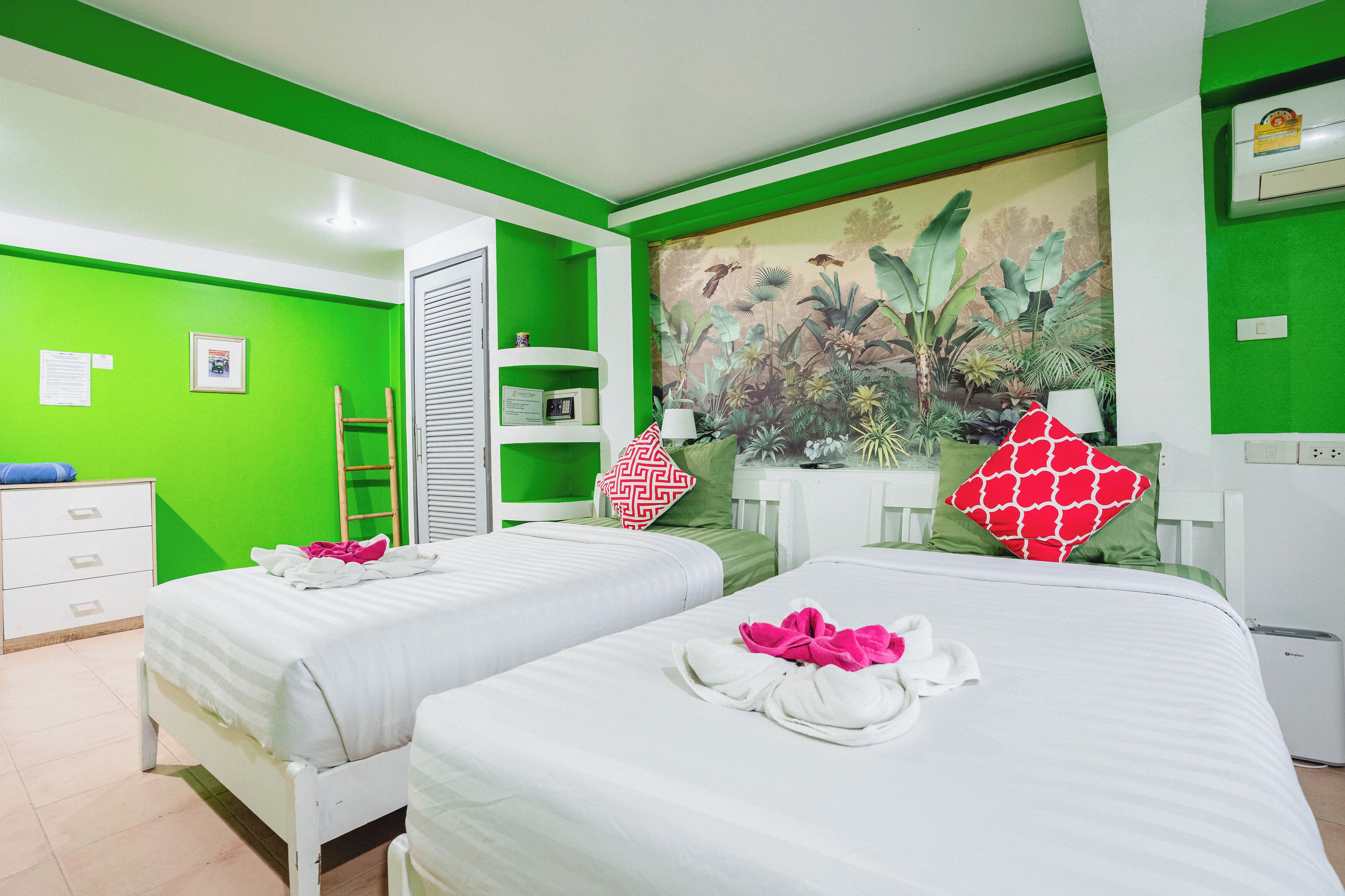 A hotel room with two beds, green walls, a tropical mural, a desk, and a white wardrobe.
