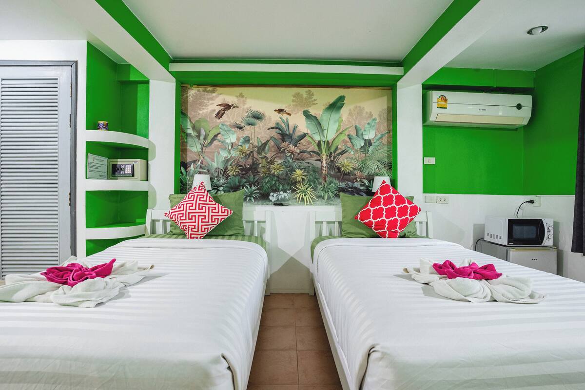 Habitación de hotel con dos camas, una pared de acento verde, un mural, un escritorio con silla, un televisor y un mueble pequeño.