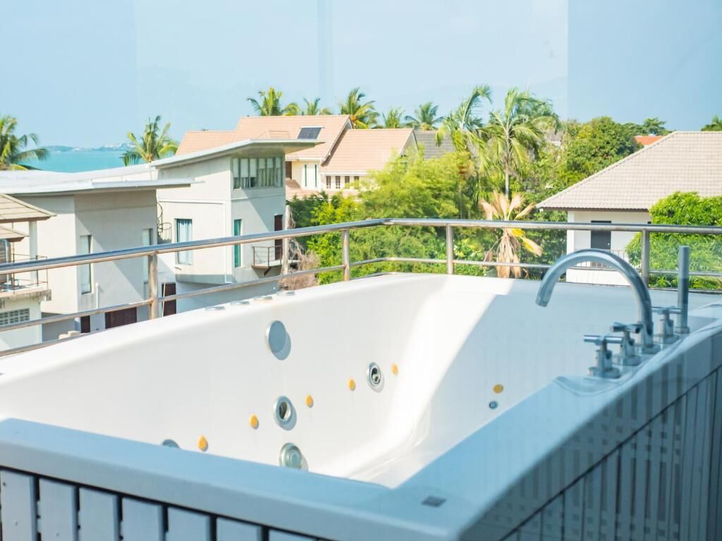 A large white hot tub with multiple jets, surrounded by a metal railing, set against a backdrop of residential buildings and palm trees.