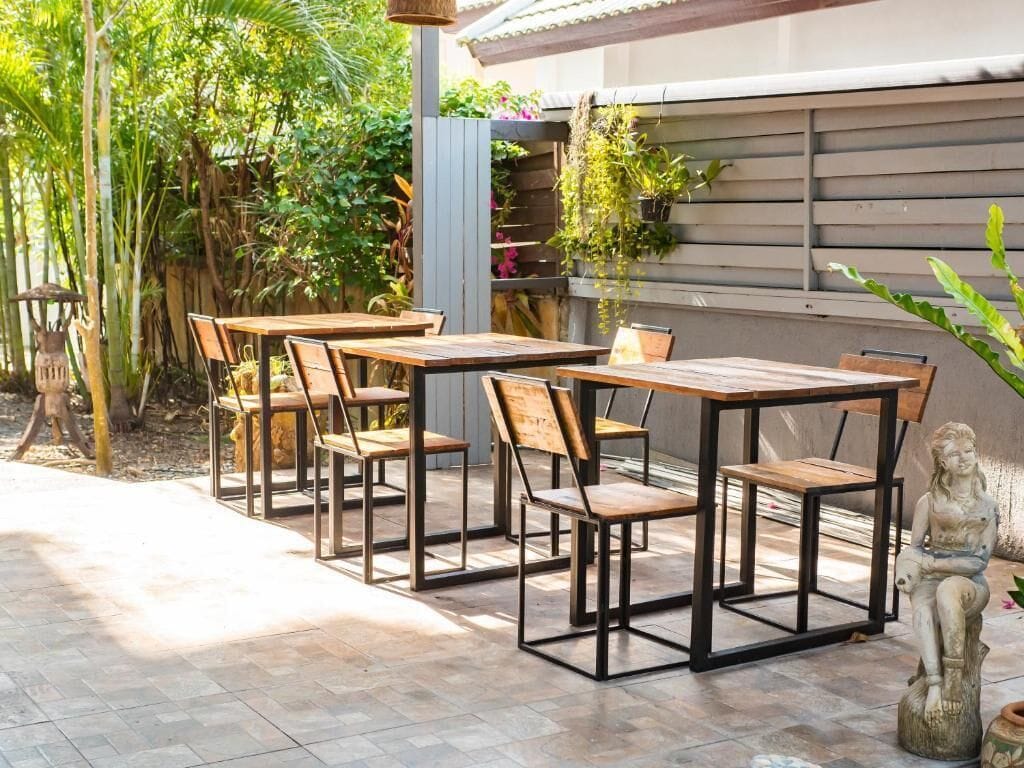 Un patio al aire libre con mesas y sillas de madera, rodeado de exuberante follaje y un suelo de piedra.