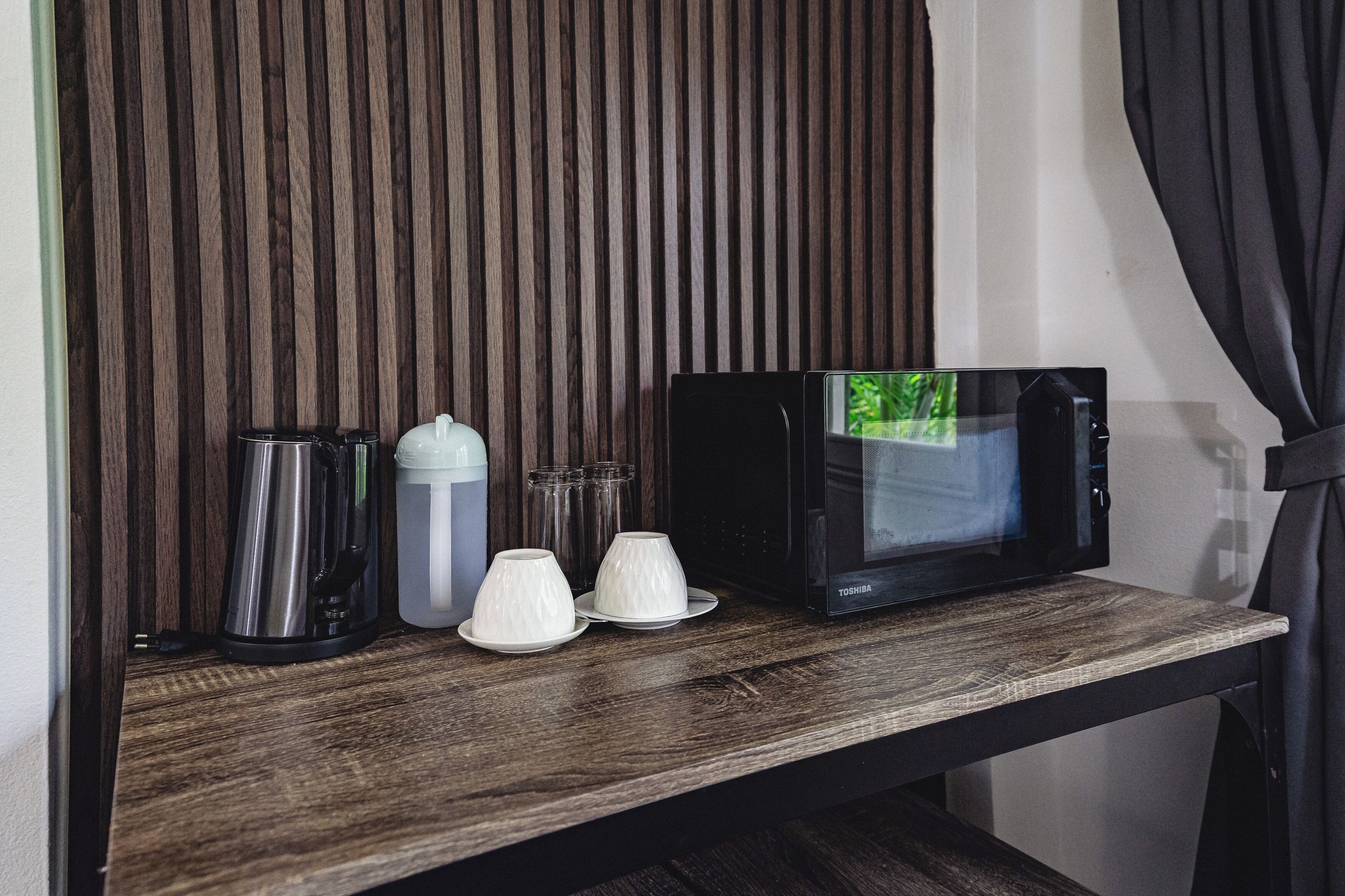 A wooden shelf with a black microwave, a white electric kettle, and two white cups.