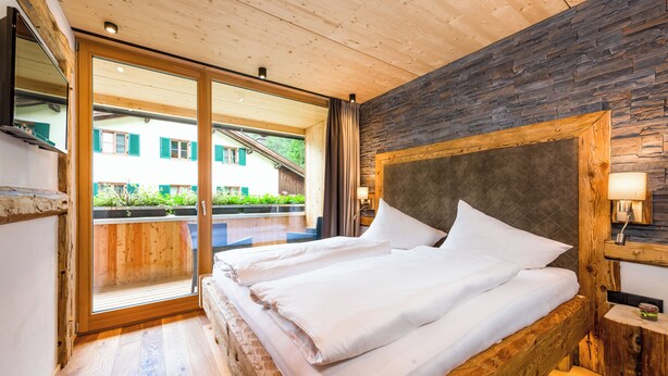 A wooden-framed bed with white bedding in a room with a stone wall and a large window. - Erholungswelt Lechrefugium (Steeg)