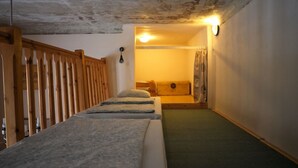 Un couloir étroit avec deux lits alignés le long du mur, une rambarde en bois d’un côté et un luminaire au plafond.