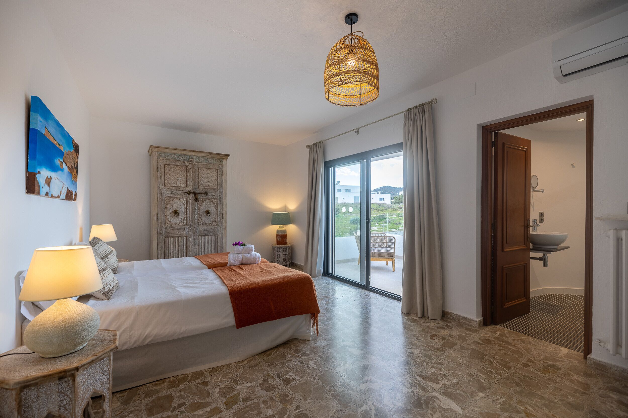 A bedroom with a bed, bedside lamps, a decorative wall piece, a wicker ceiling light, a wardrobe, and a bathroom with a sink and shower.