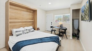 A bedroom with a large bed, a small table with chairs, a door, and a window with blinds.