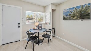 A room with a dining table and chairs, a door, a window with blinds, and a framed picture on the wall.
