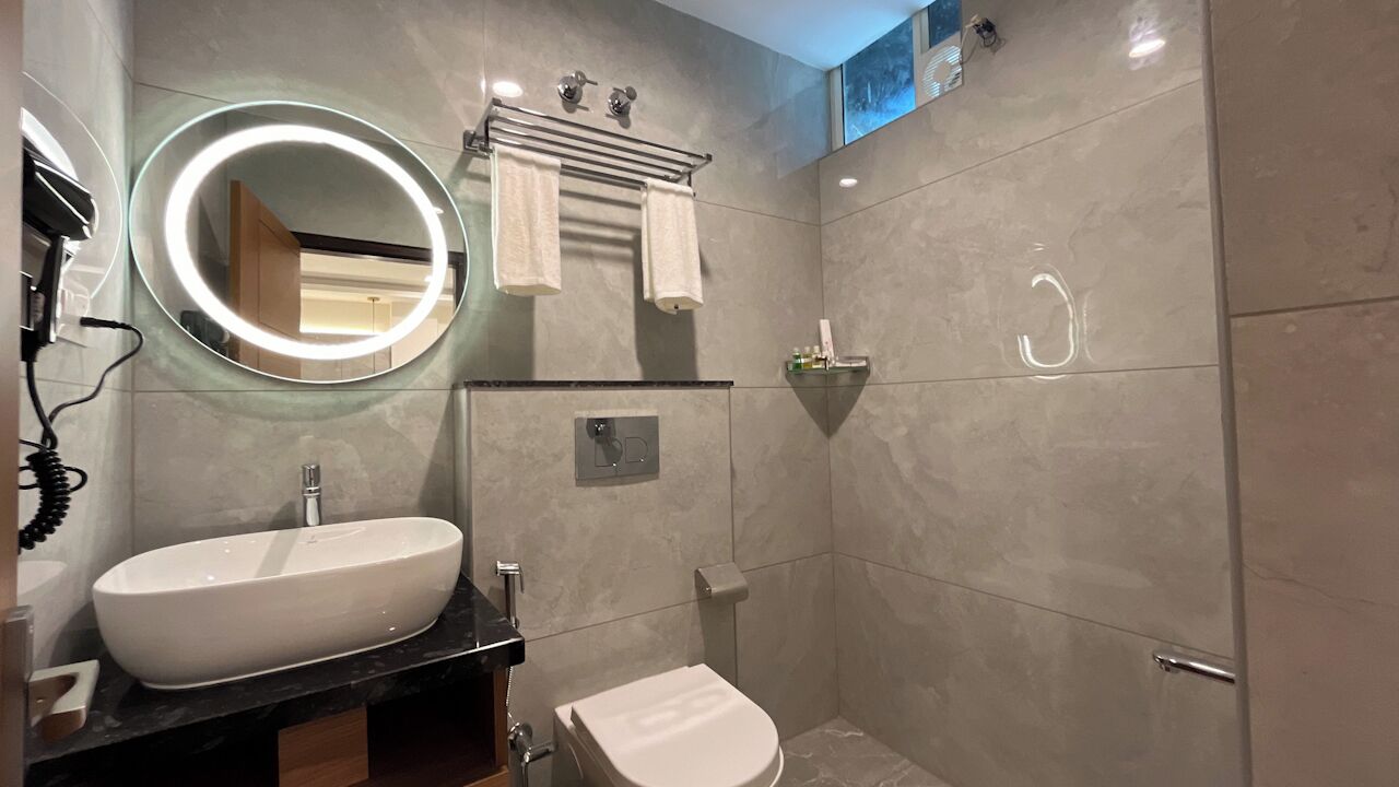 Et moderne badeværelse med en stor, oval håndvask, et rundt spejl med lys, et toilet og et bruseområde.