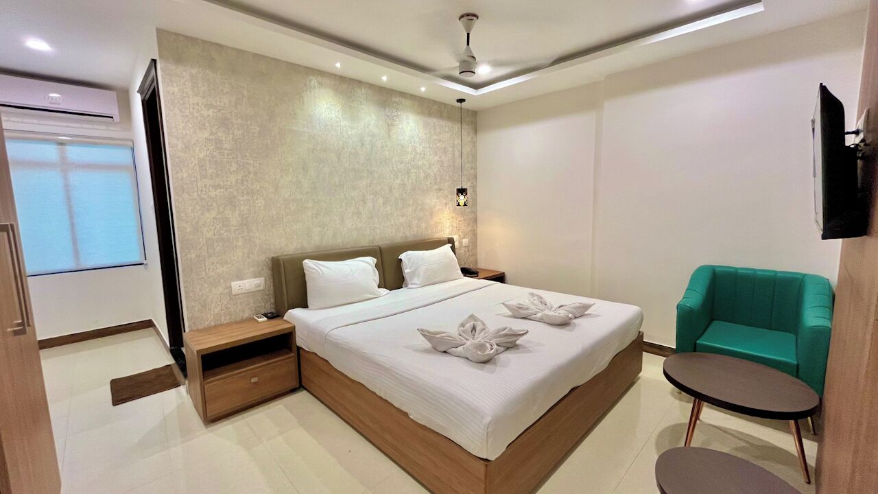 Ein modernes Hotelzimmer mit einem großen Bett, einem kleinen runden Tisch, einem petrolfarbenen Sessel und einem flachbildfernseher an der Wand.