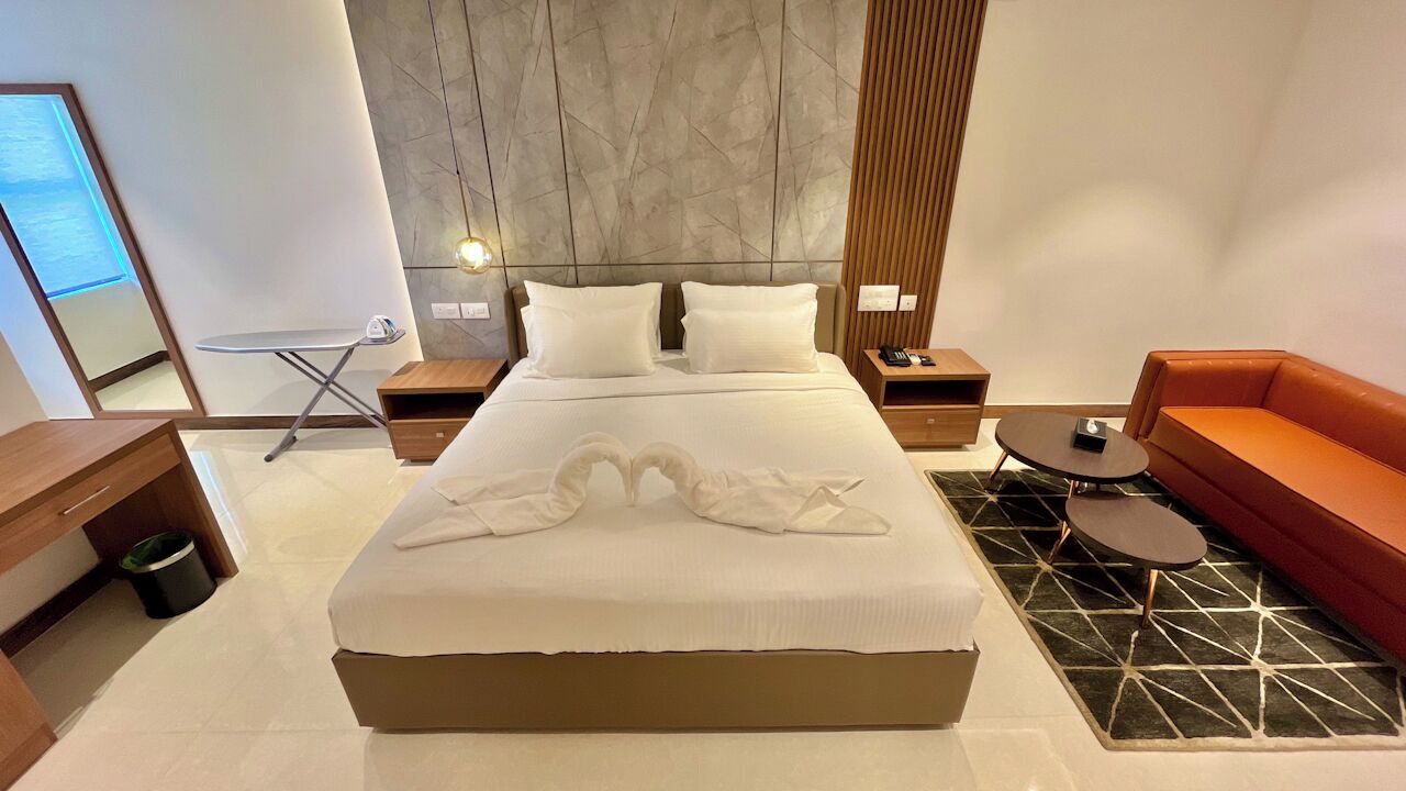 A modern hotel room with a large bed, a wooden headboard, a red sofa, a small round table, and a mirror.