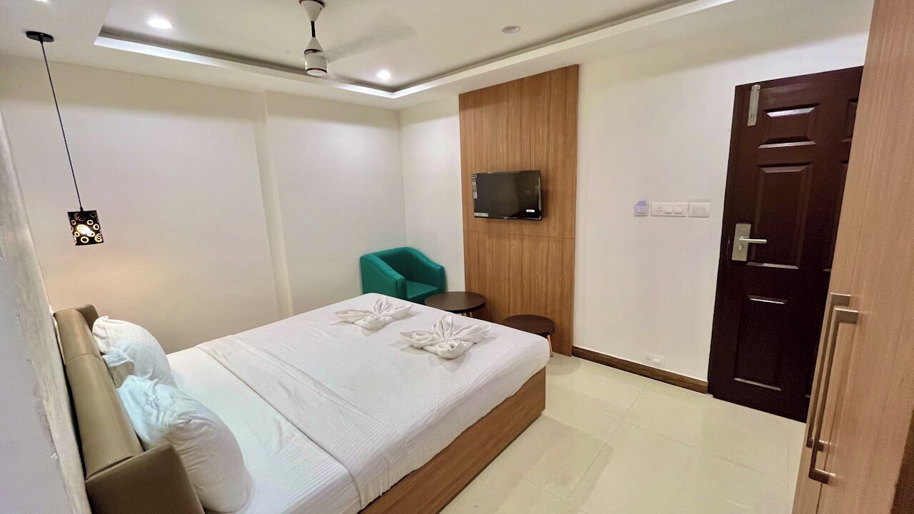 A hotel room with a bed, a teal armchair, a TV mounted on the wall, and a wooden door.