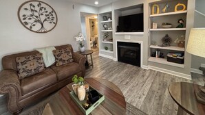Ein Wohnzimmer mit braunem Ledersofa, einem Couchtisch mit Dekoration, einem Kamin, einem Fernseher und einem Deckenventilator.