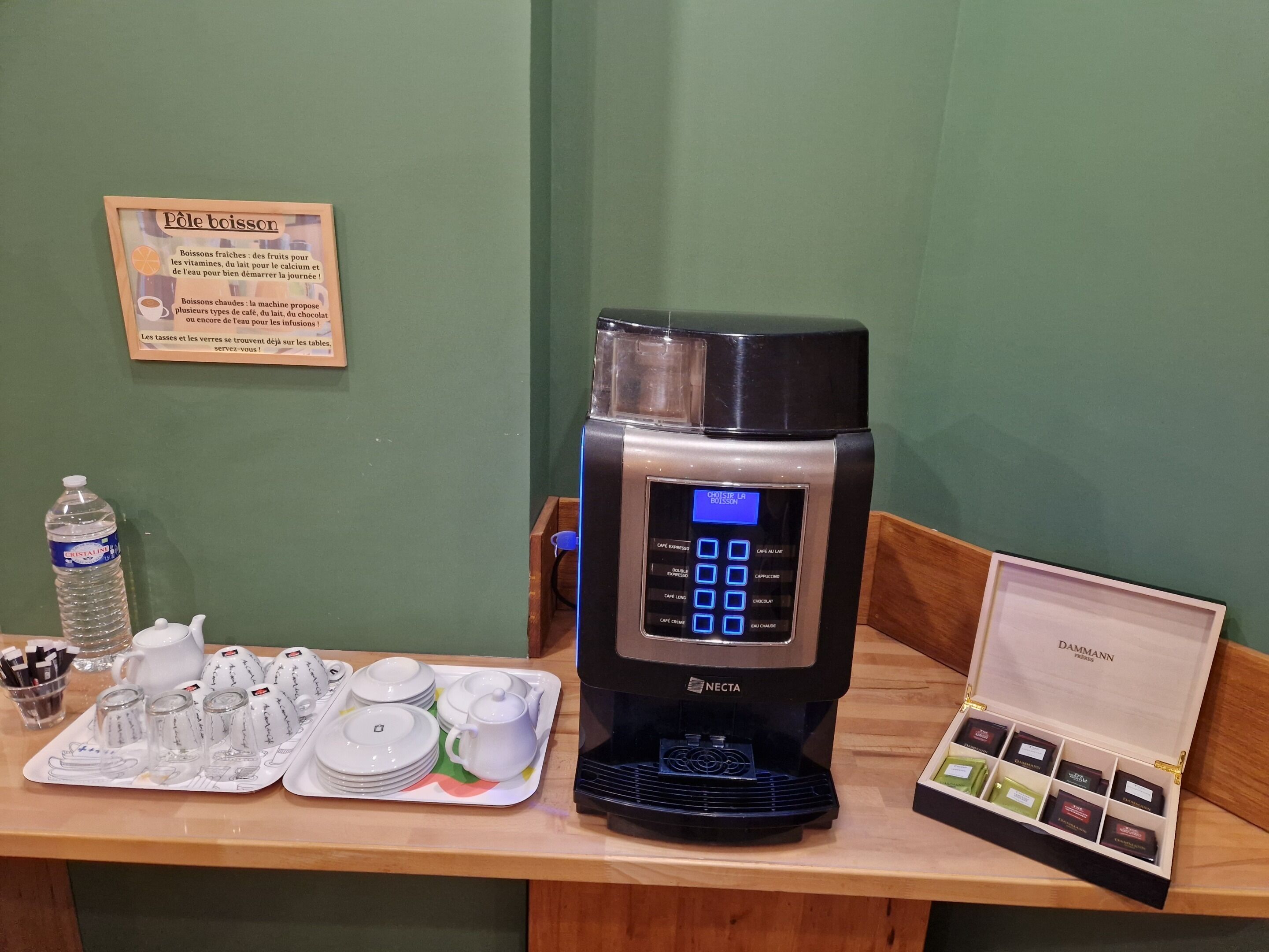 Une machine à café dotée d’un écran numérique, un plateau avec des tasses et des soucoupes, une bouteille d’eau et une boîte de chocolats.