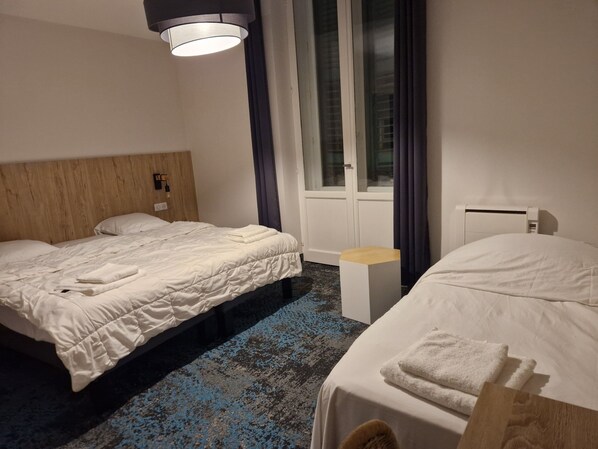 Habitación de hotel con dos camas, cabecero de madera, mesita de noche pequeña, ventana con cortinas e iluminación de techo.