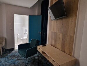 Een moderne hotelkamer met een houten vloer, een groene stoel, een houten console en een televisie die aan de muur is bevestigd. Een badkamer is zichtbaar door een geopende deur.