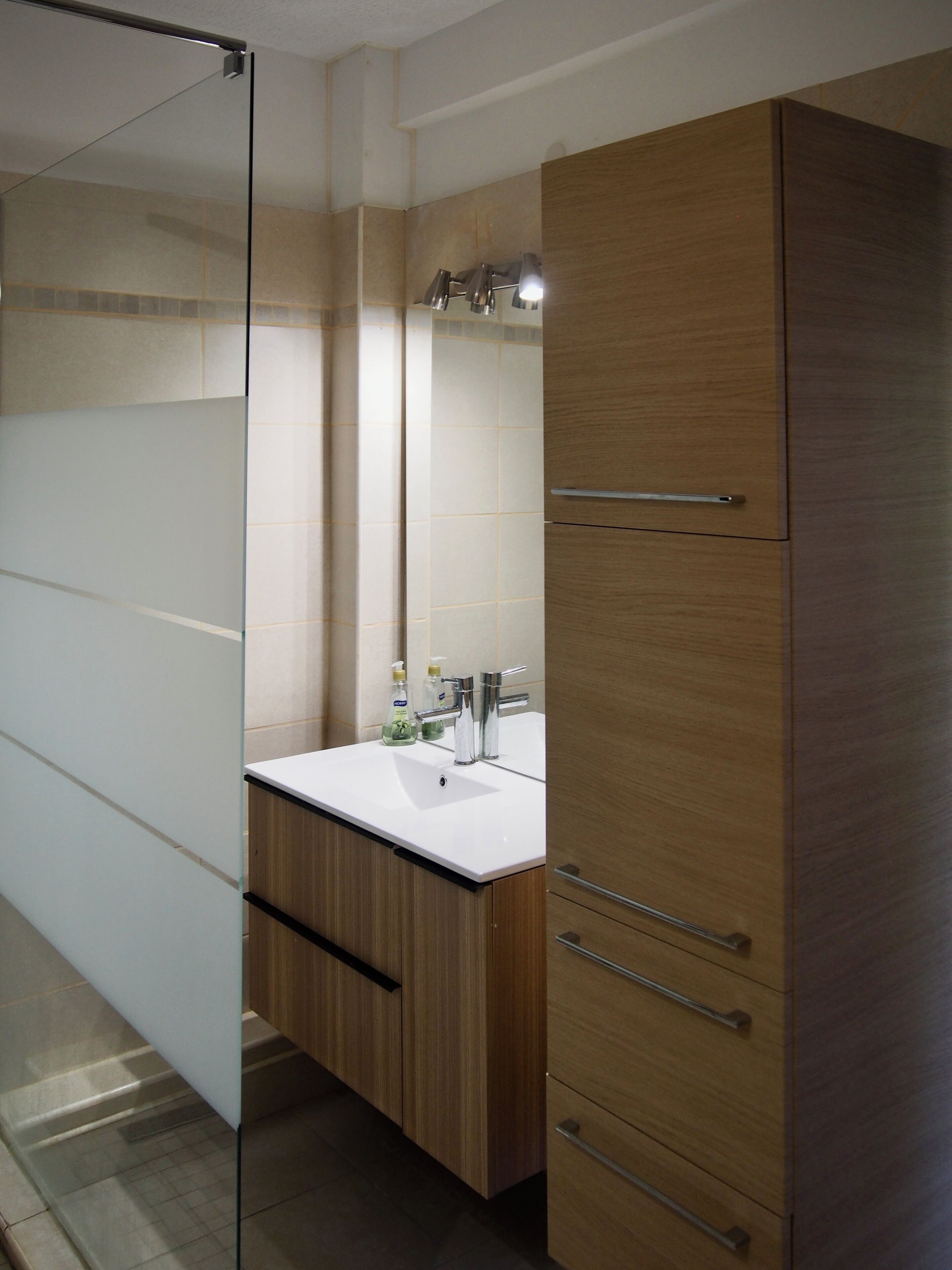 Une salle de bain moderne avec une douche à parois de verre, un meuble-lavabo en bois doté d’un plan de travail blanc et un miroir au-dessus du lavabo.