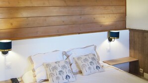 Ein Schlafzimmer mit Holz-Kopfstück, zwei Nachttischlampen, einem Bett mit weißer Bettwäsche und zwei Zierkissen mit Palmenmotiven.