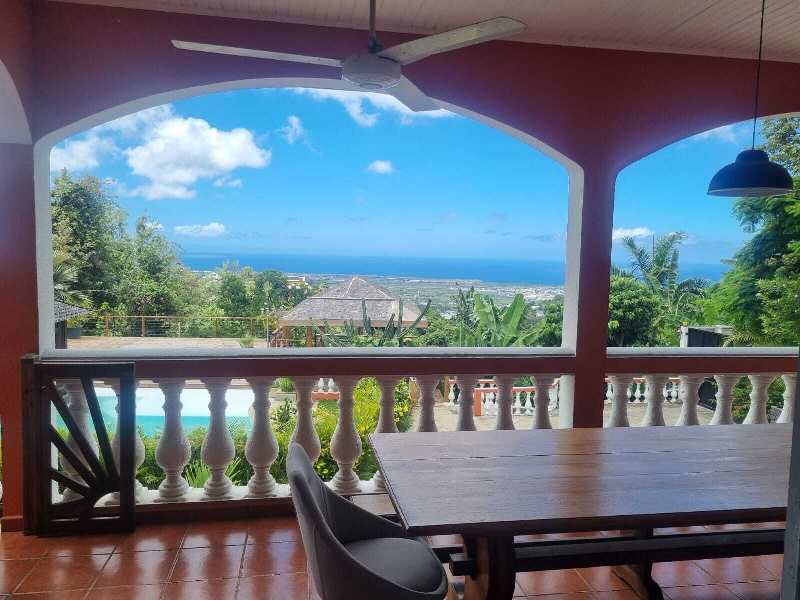 Ein überdachter Außenbereich mit Tisch und Stühlen, einem Deckenventilator und Blick auf das Meer und die grüne Landschaft.