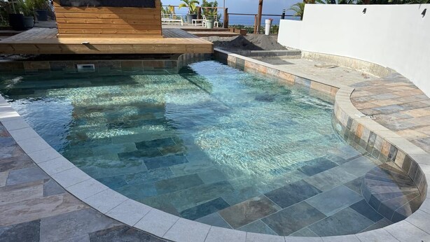 Ein Pool mit geschwungener Kante, umgeben von Steinfliesen und einer Holzterrasse.