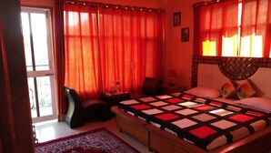 A bedroom with a large bed, a nightstand, a chair, and a window with red curtains. - Love Kush Cottage  (Taradevi)