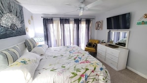 A bedroom with a bed, a ceiling fan, a TV, a dresser, and a window with curtains. - Tradewinds 151 1 Bedroom Condo (St. Augustine)