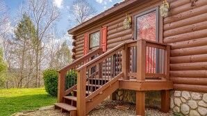 A wooden log cabin with a wooden staircase leading to a deck, surrounded by a stone foundation and a grassy yard. - Greenbrier Woods 2 Bedroom Cabin (Gatlinburg)