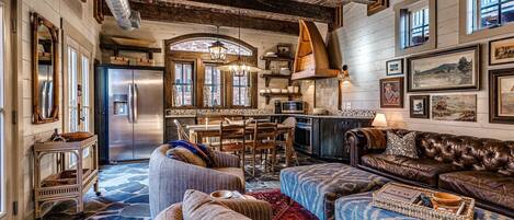Uma sala de estar ampla com um sofá grande, uma mesa de centro e uma lareira. Há uma cozinha com geladeira, micro-ondas e armários de madeira. O ambiente possui teto de madeira rústica e piso de pedra.