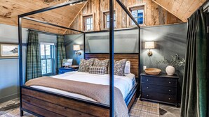 Een vierposterbed in een rustieke kamer met houten balken, een kroonluchter en uitzicht op de omgeving door ramen.