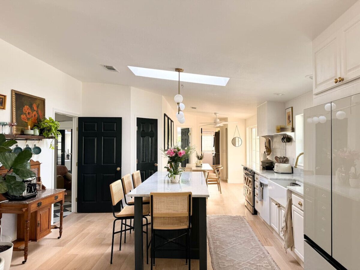A modern kitchen with a central island, wooden chairs, a dining table, and a black door.