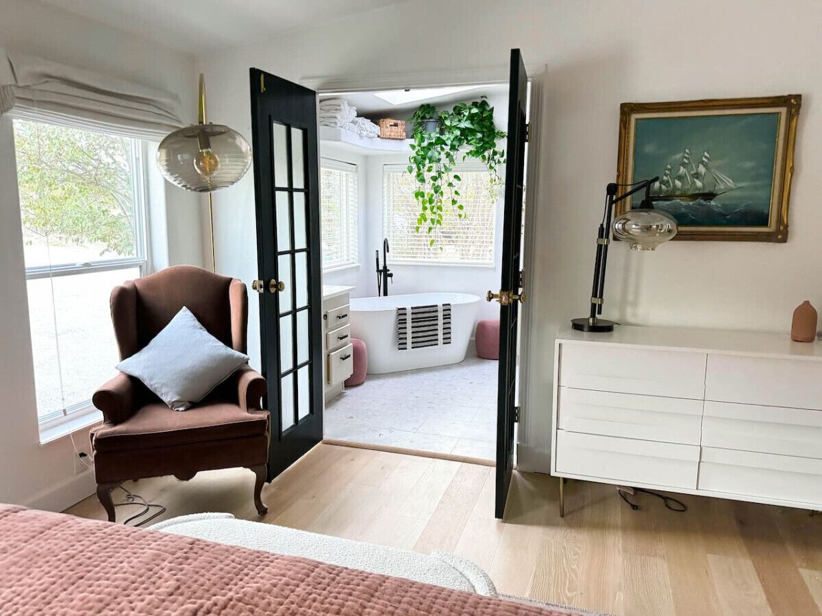 A room with a brown armchair, a white dresser, a painting, and a window with a view of a garden.