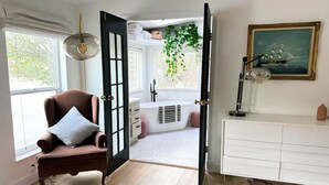 Una habitación con un sillón marrón, un tocador blanco, un cuadro y una ventana con vistas a un jardín.