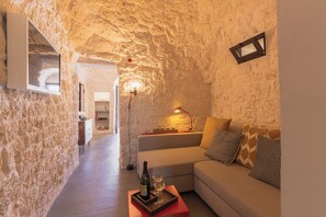 Una habitación de paredes de piedra con un sofá, una pequeña mesa con una botella y un vaso, y un televisor instalado en la pared.
