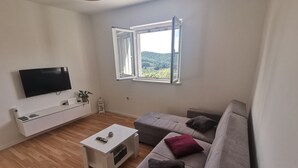 A modern living room with a flat-screen TV, a white coffee table, a grey sofa with cushions, and a window with a view of greenery. - Perfect Getaway Near Korčulas Scenic Sights and Beaches (Korcula)