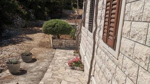 A stone pathway leading to a building with a stone facade and wooden shutters. There are potted plants and a small tree along the path.