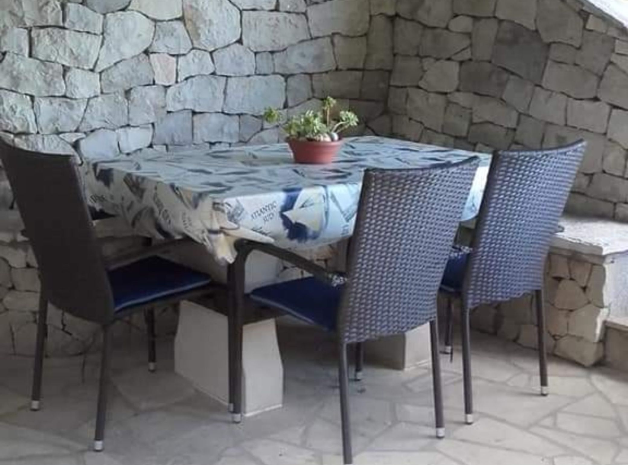 A stone wall, a round table with a patterned tablecloth, four chairs, and a potted plant.