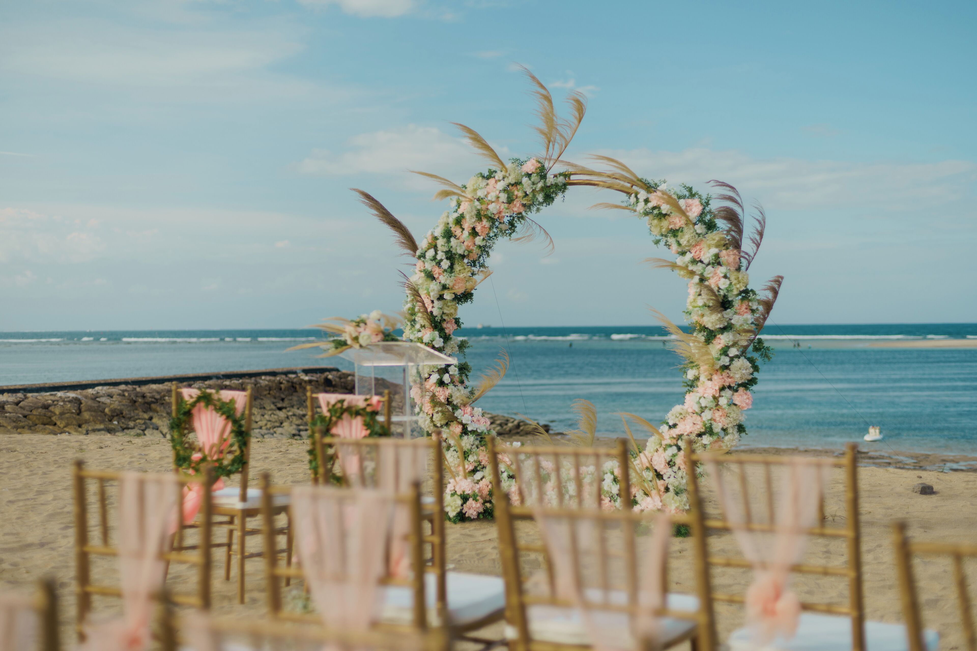 A beach wedding setup with a floral arch and wooden chairs.