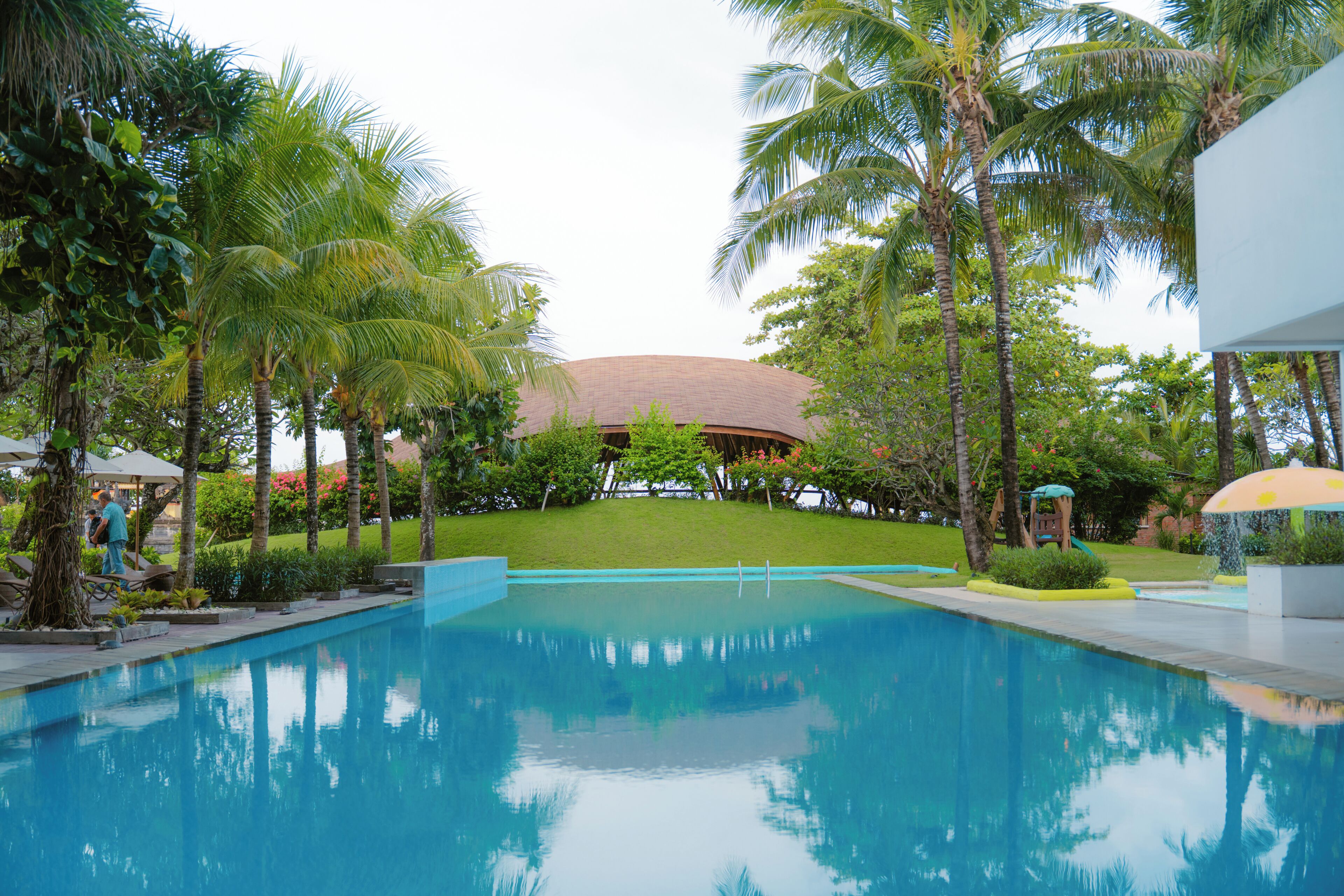 Una piscina con superficie de agua azul cristalina, rodeada de palmeras y un edificio con techo curvo.