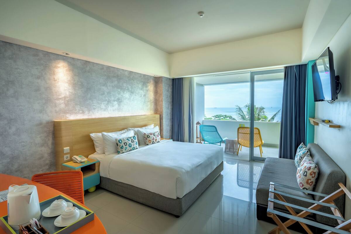 A hotel room with a large bed, a sofa, a TV, a balcony with a view, and a table with a tray of items.