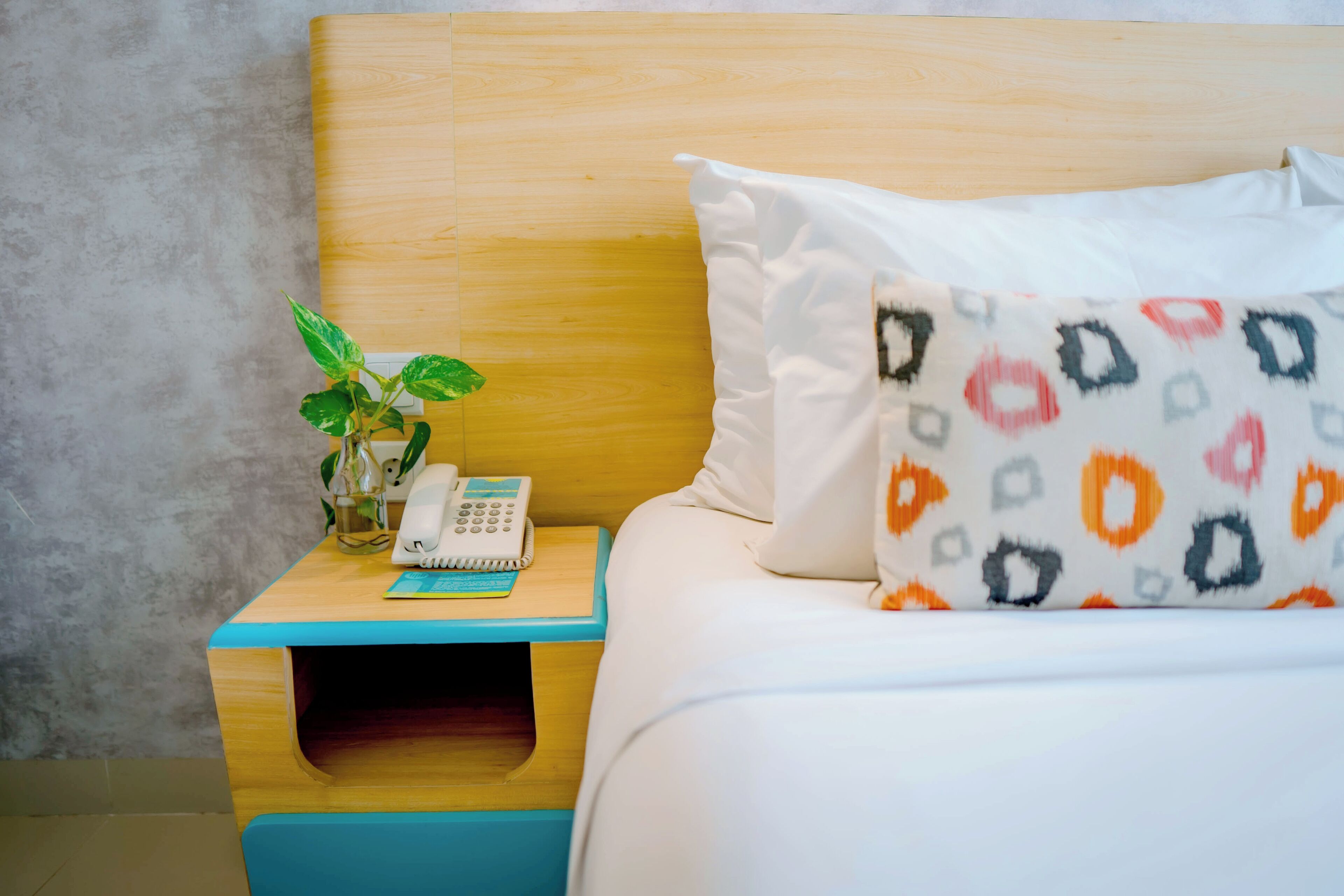 A neatly made bed with a patterned pillow, a bedside phone, a small plant, and a wooden nightstand with a blue accent.