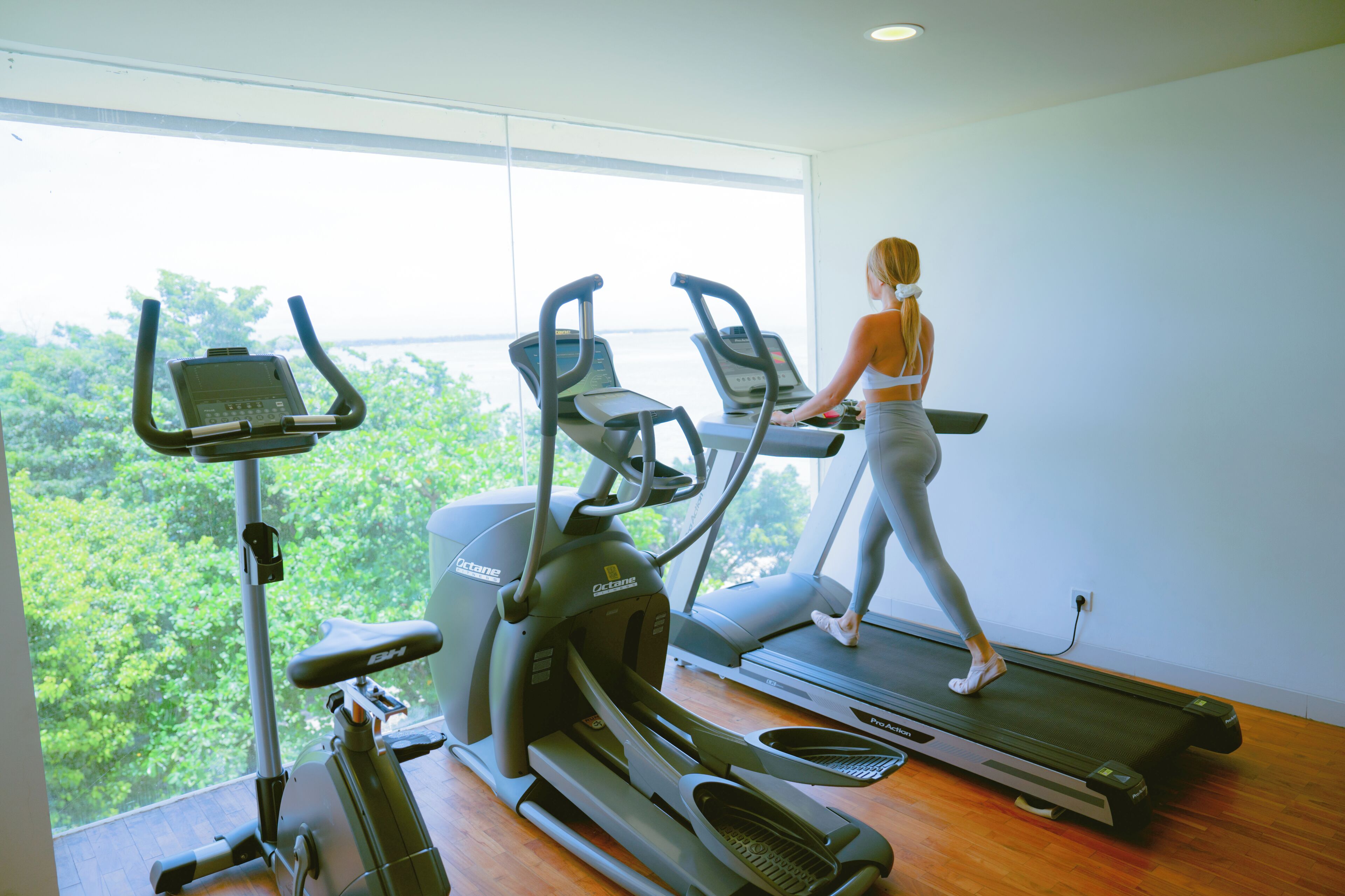 A woman walking on a treadmill in a room with large windows and a stationary bike.