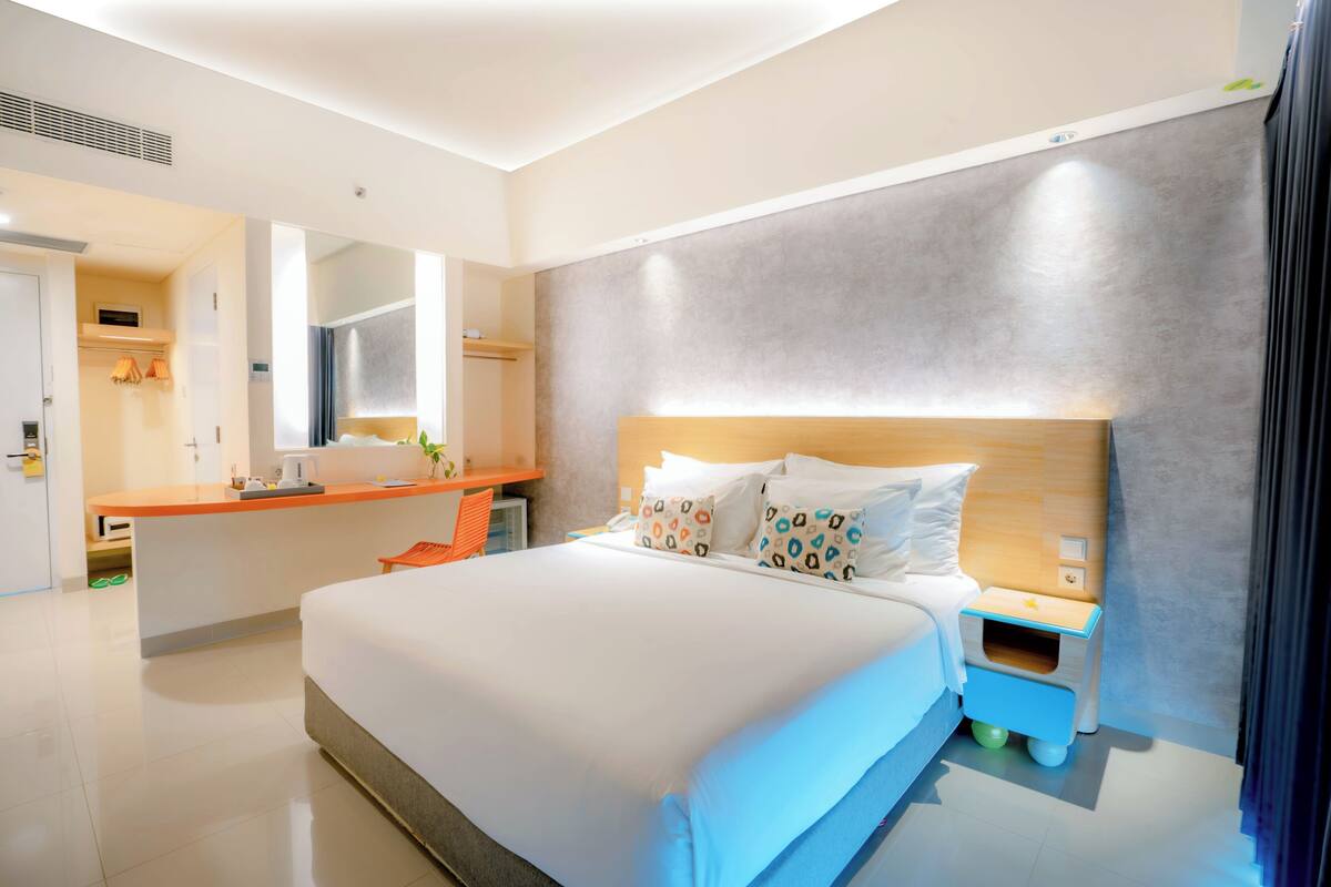 A modern hotel room with a large bed, a bedside table, a small wooden nightstand, a built-in desk with a chair, and a large mirror.