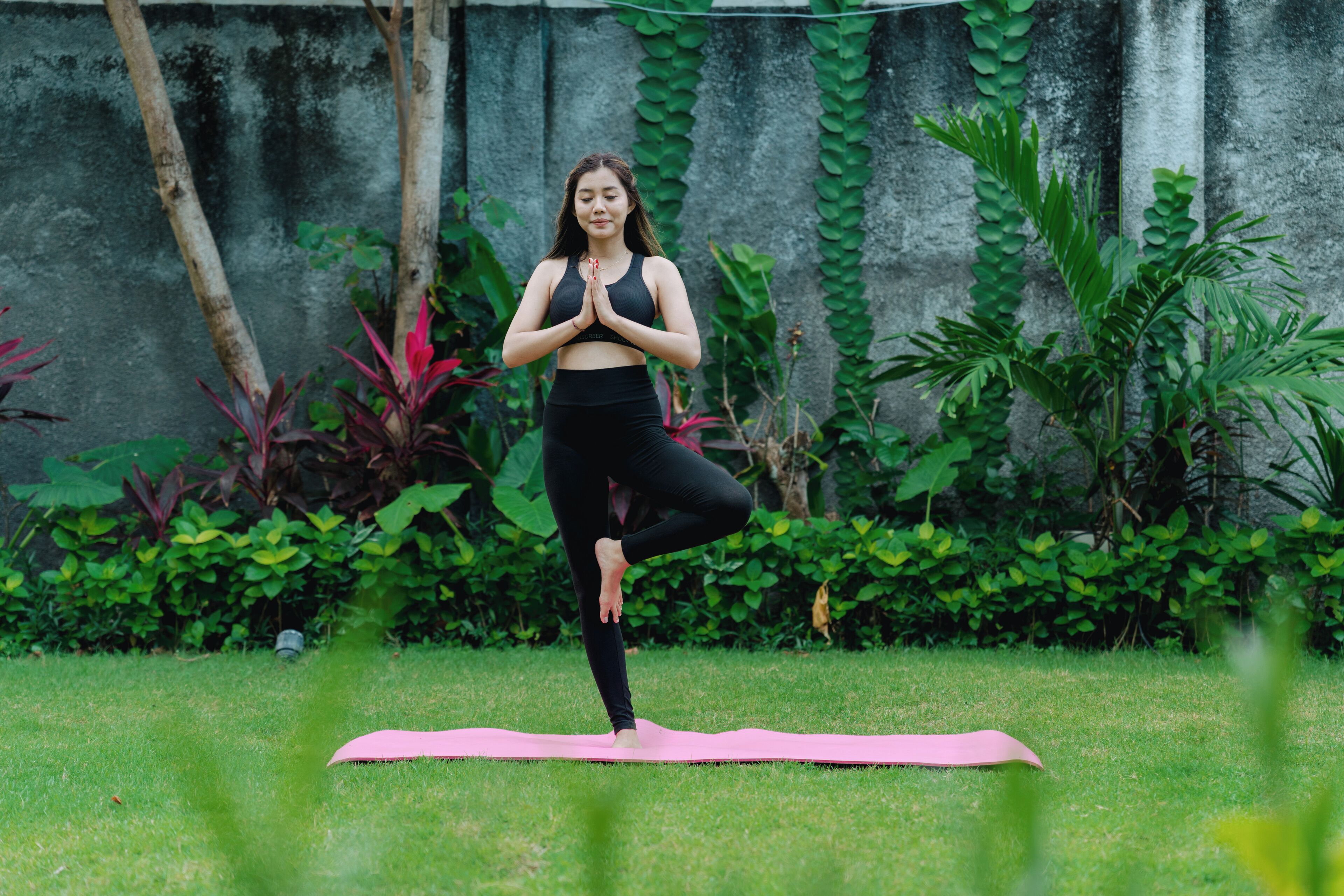 A person performing a yoga pose on a pink mat in an outdoor setting with greenery and a concrete wall in the background.