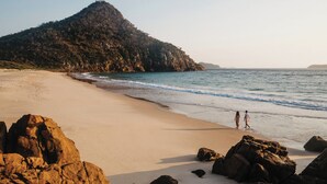 Uma praia com formações rochosas e uma grande colina ao fundo.