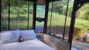 Une chambre à coucher avec un lit, une table de chevet et une vue sur un jardin luxuriant, visible à travers de grandes fenêtres.