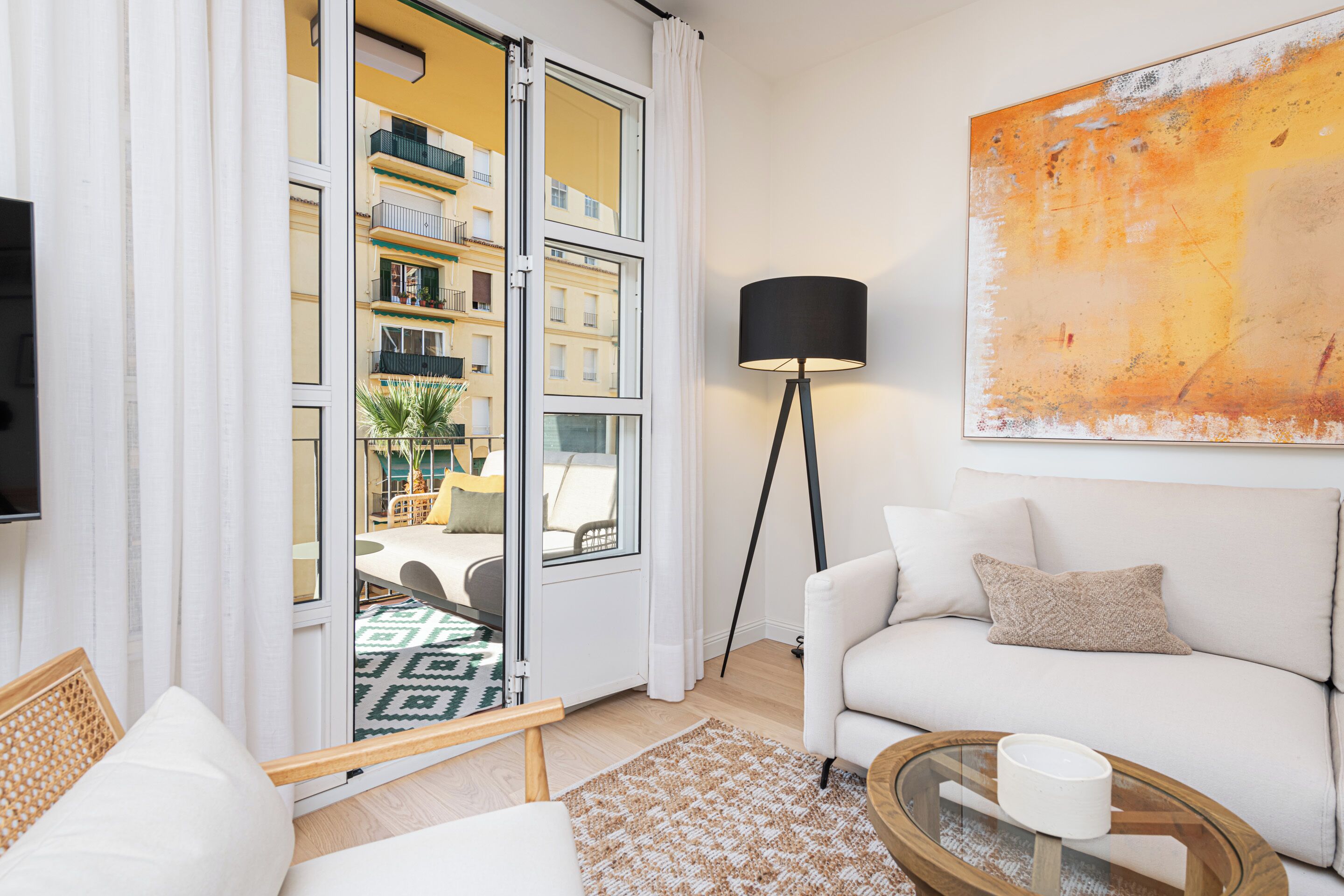 A modern living room with a white sofa, a glass coffee table, a floor lamp, and a view of a balcony with outdoor furniture.