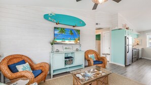 A living room with wicker chairs, a glass coffee table, a teal surfboard, and a beach-themed painting. - Adorable Port Aransas beach cottage with community pool and outdoor spaces.  Clo (Port Aransas)