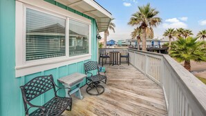 A wooden deck with a covered seating area, a large window, and a view of palm trees and other buildings. - Adorable Port Aransas beach cottage with community pool and outdoor spaces.  Clo (Port Aransas)