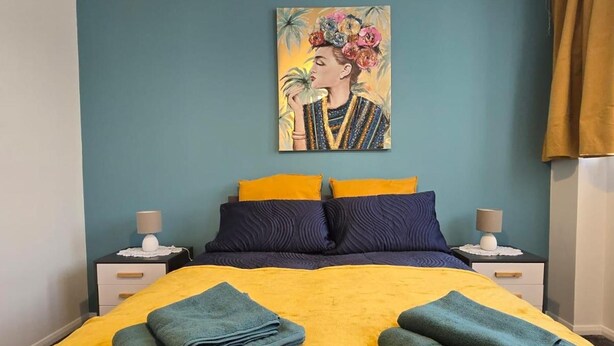 Ein Bett mit gelber Bettdecke und zwei gefalteten Handtüchern, ein Bild an der Wand sowie zwei Nachttische mit Lampen.