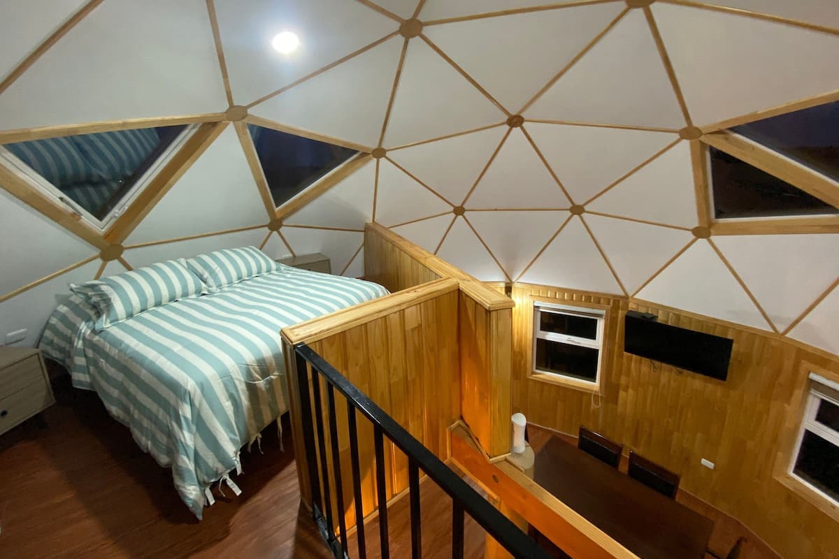 A room with a geometric ceiling design, a bed with striped bedding, a wooden table, and a small window.