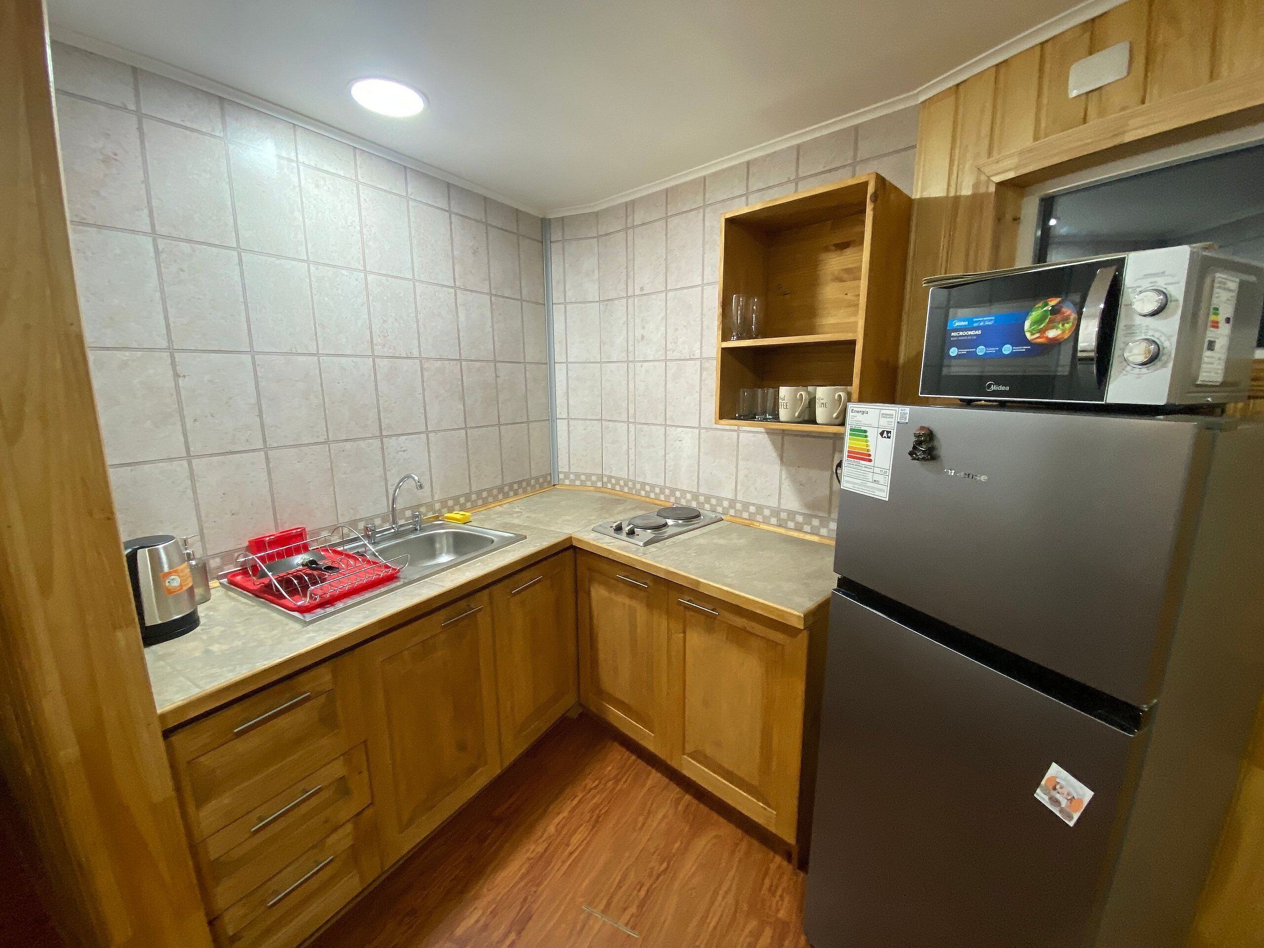 Una cocina compacta con gabinetes de madera, un lavamanos de acero inoxidable, un microondas y un refrigerador.
